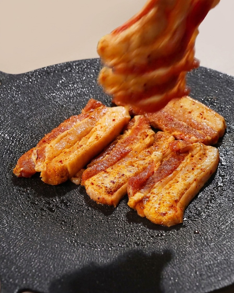 Several strips of seasoned pork belly being organized on a hot grill pan with a hand visible at the top.