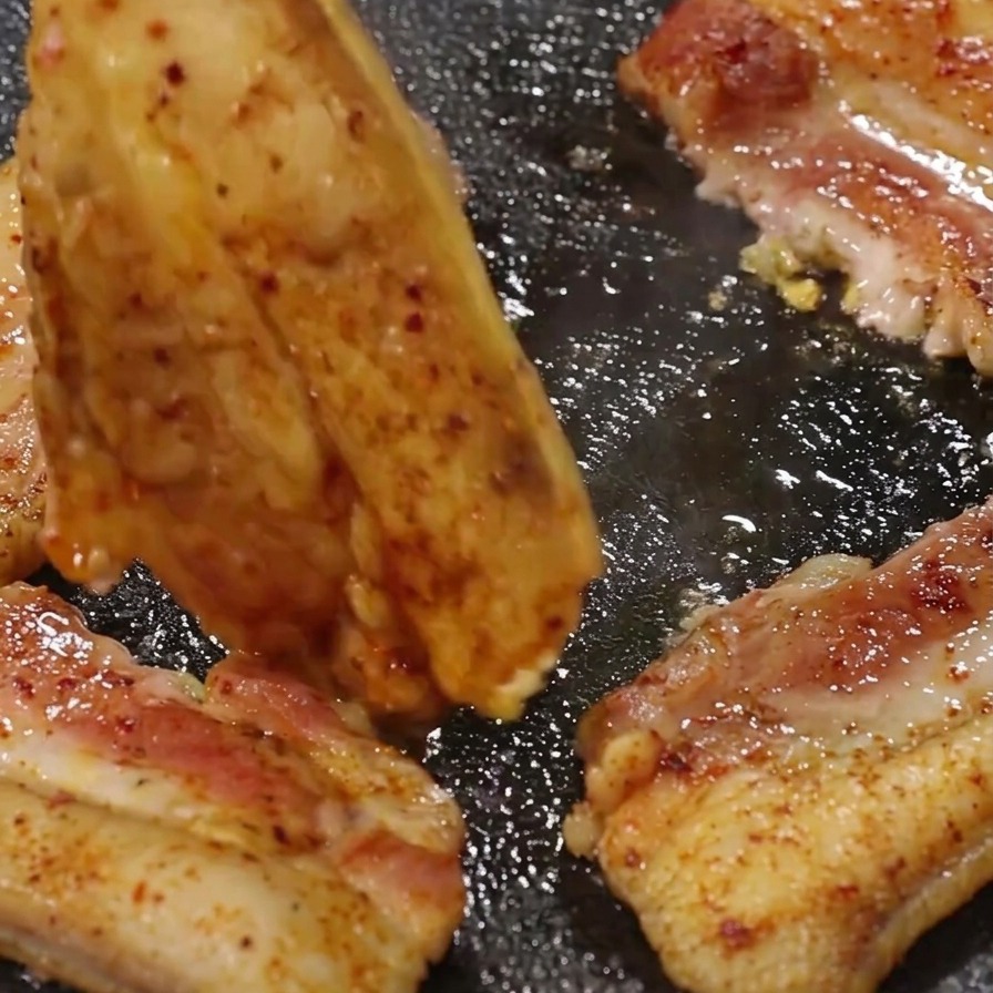 Metal tongs flipping a piece of browning pork belly on a hot, sizzling grill pan.