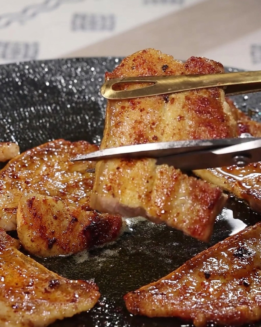 Metal kitchen scissors cutting through a piece of grilled pork belly held by tongs over a sizzling pan.