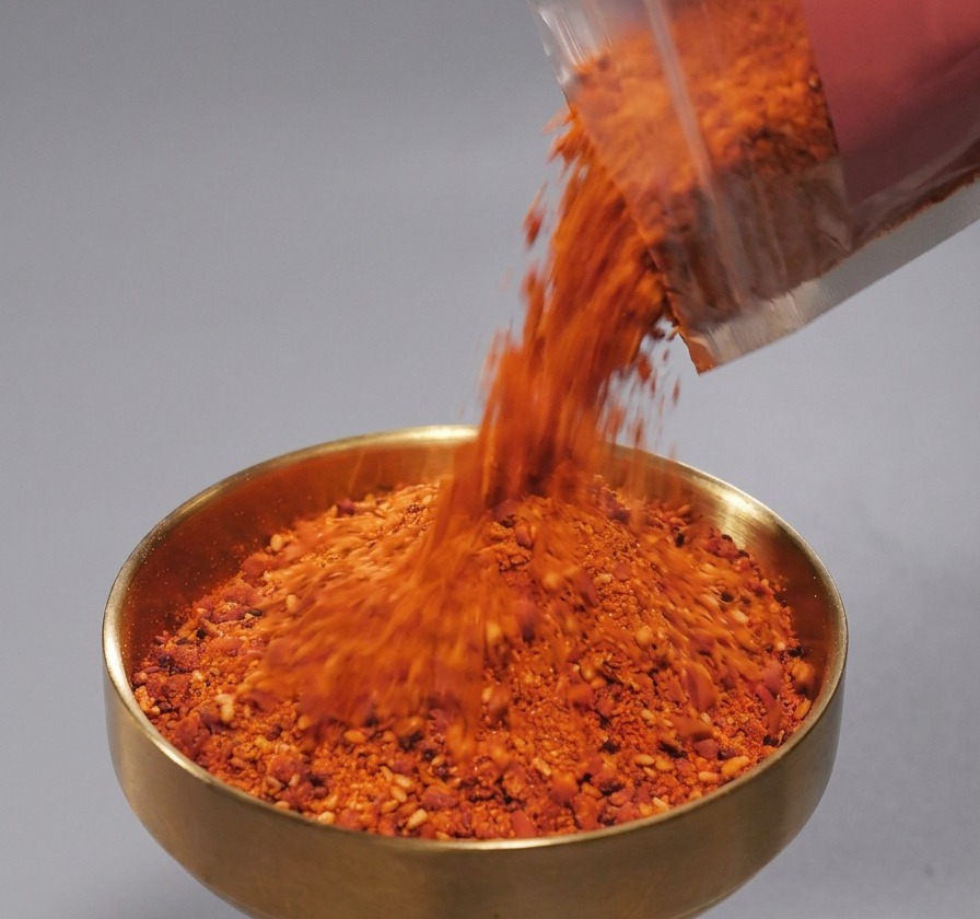 Red spice powder with sesame seeds being poured from a plastic packet into a brass-colored bowl.