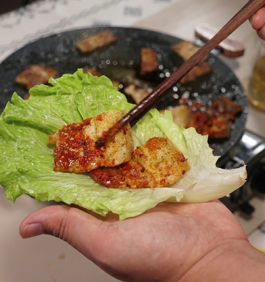 A hand holding a large green lettuce leaf while placing a piece of grilled pork belly onto it with chopsticks.