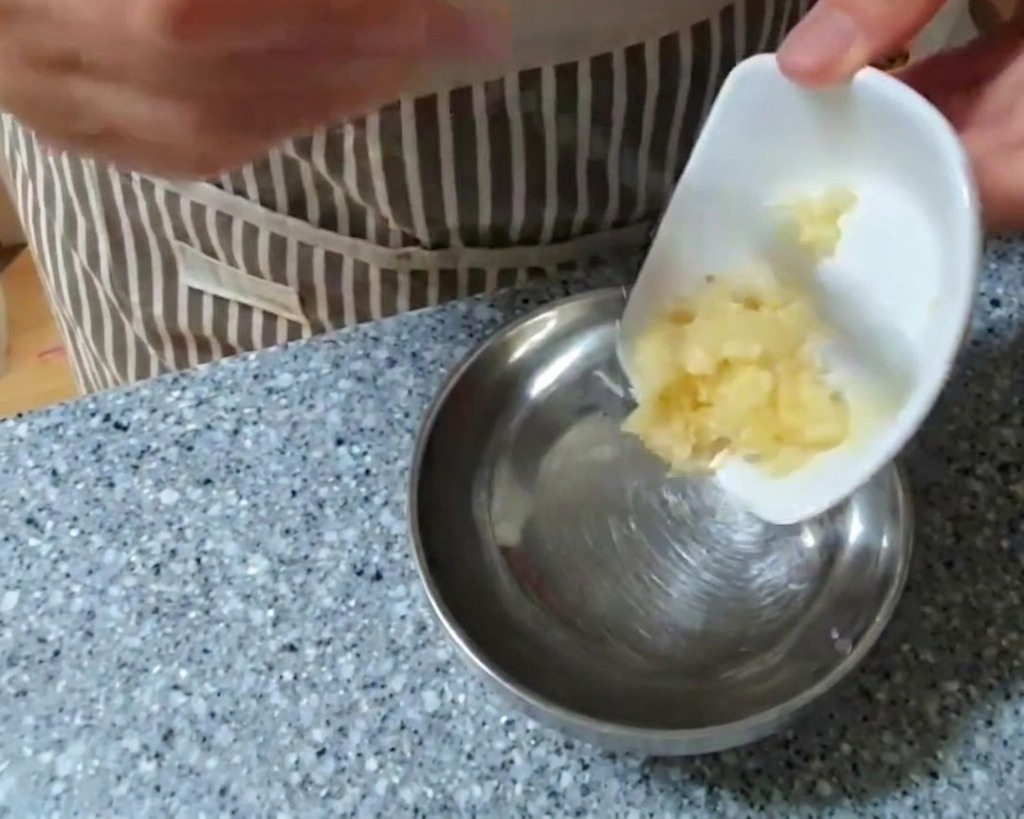 A person adding a portion of minced garlic from a small white dish into a larger stainless steel mixing bowl.