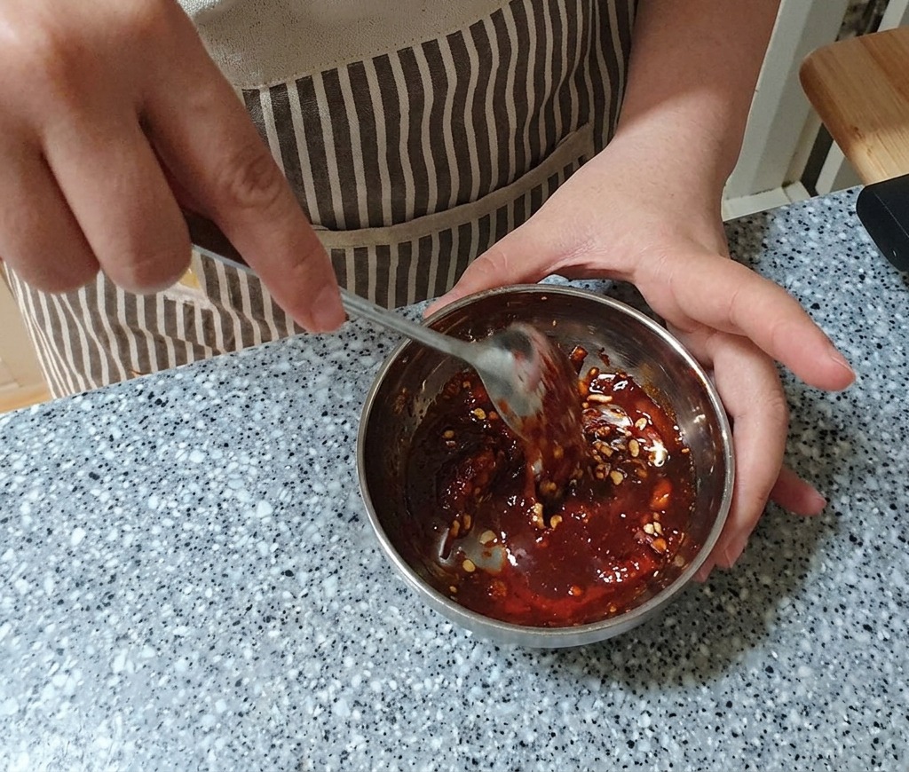 Stirring a deep red, thick marinade in a metal bowl with a stainless steel spoon until fully combined.