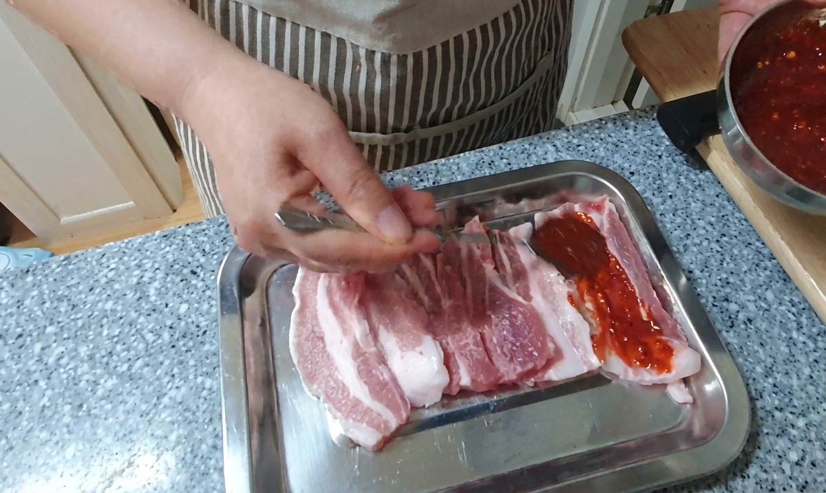 A hand using a spoon to coat strips of raw pork belly with a thick, vibrant red chili marinade on a silver tray.
