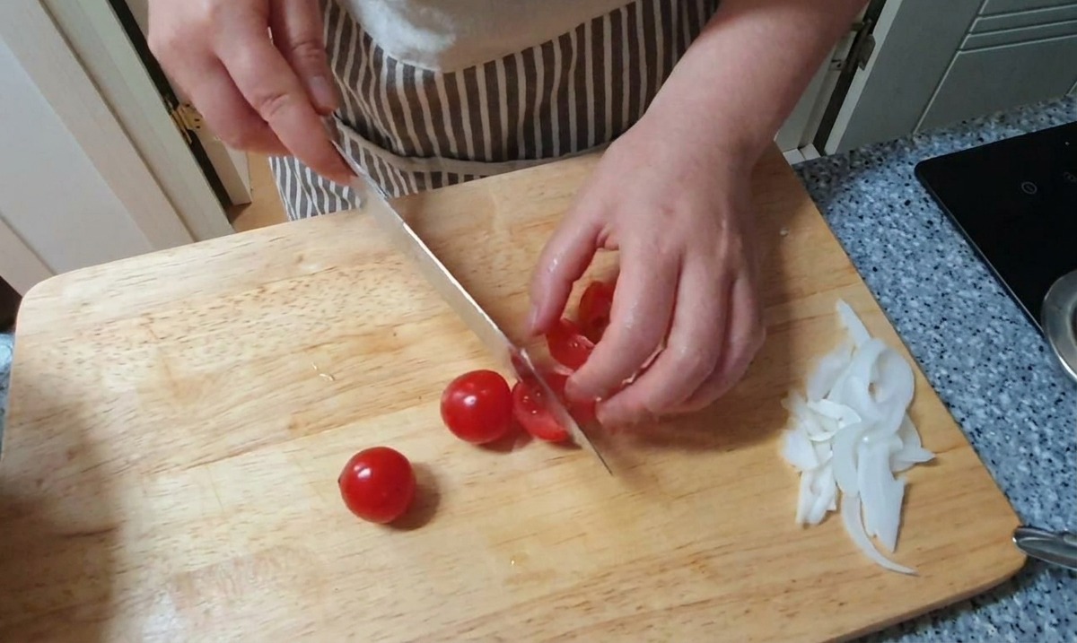 Slicing small red cherry tomatoes in half on a wooden board next to a pile of sliced white onions.
