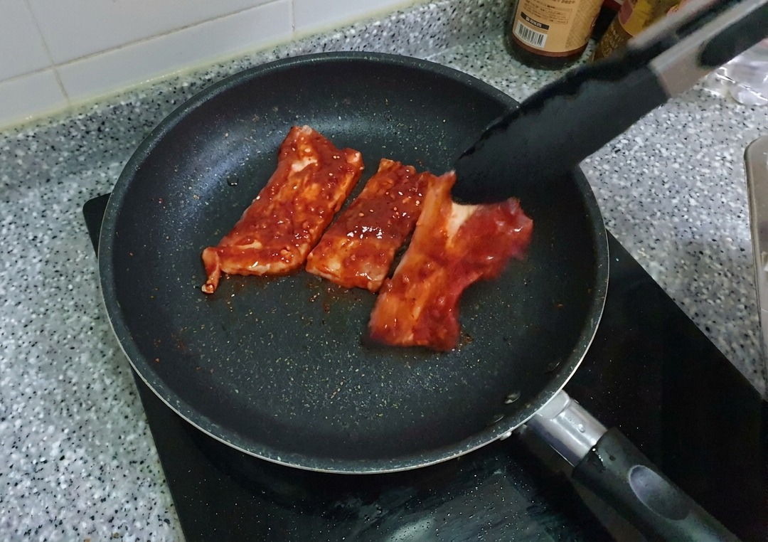 Marinated red pork belly strips being placed into a black non-stick frying pan using tongs.