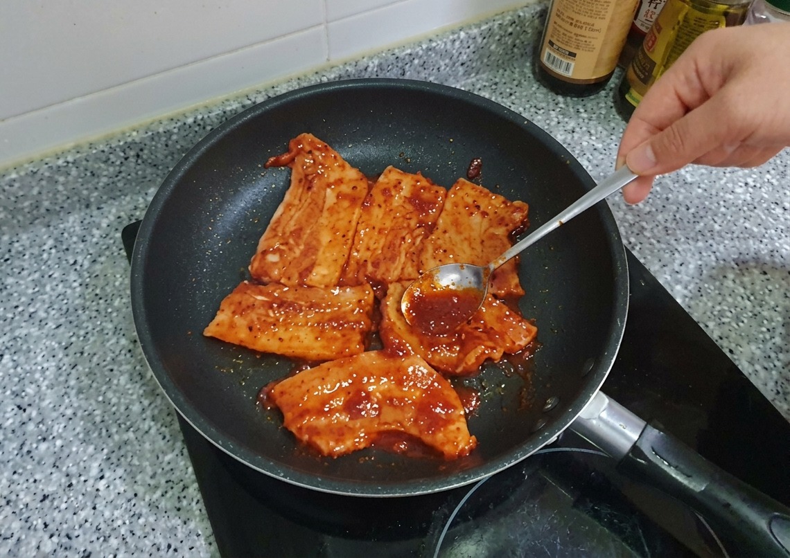 A spoon spreading thick red Gochujang marinade over pork belly slices in a pan.