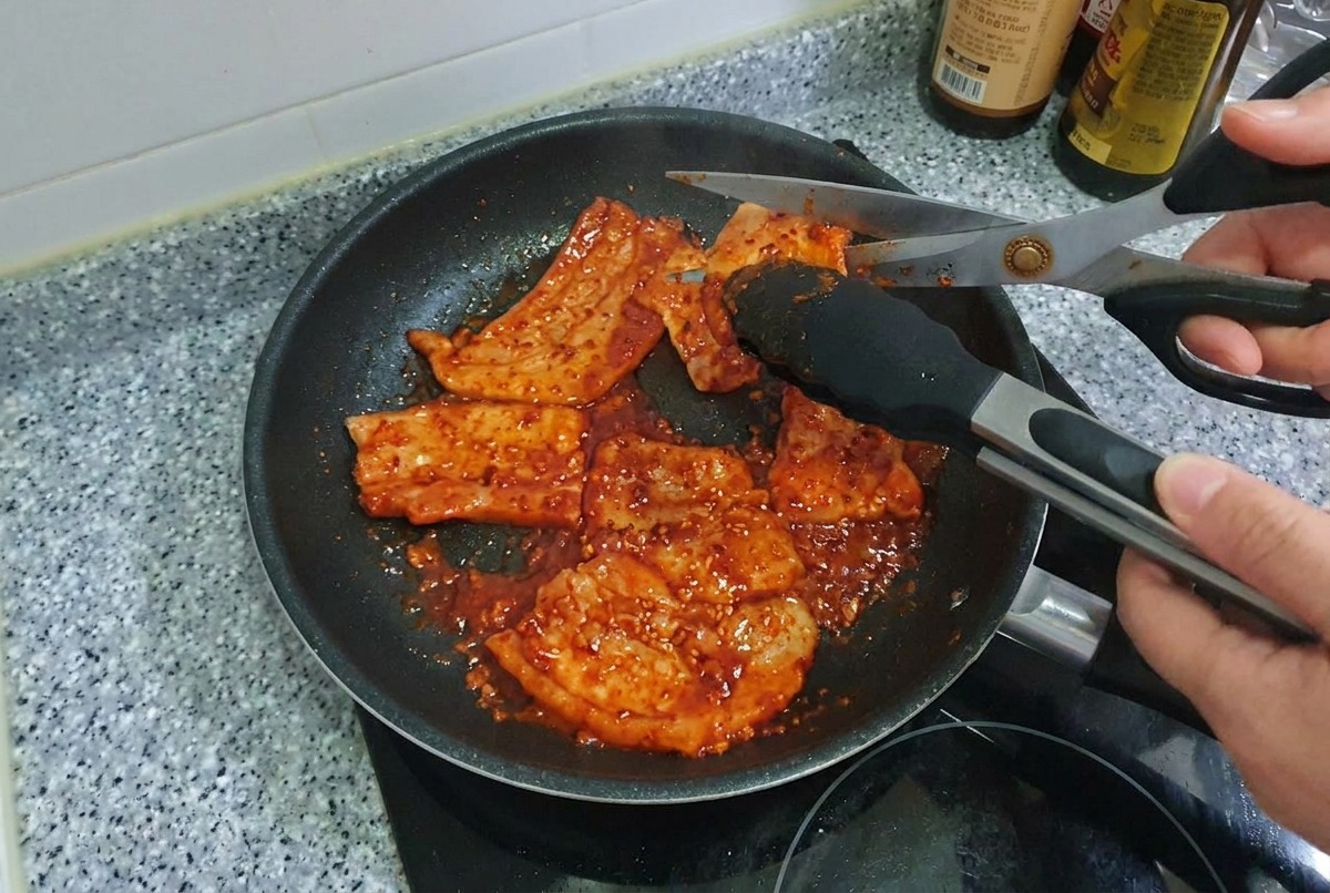 Kitchen scissors cutting through strips of spicy grilled pork belly inside a frying pan.
