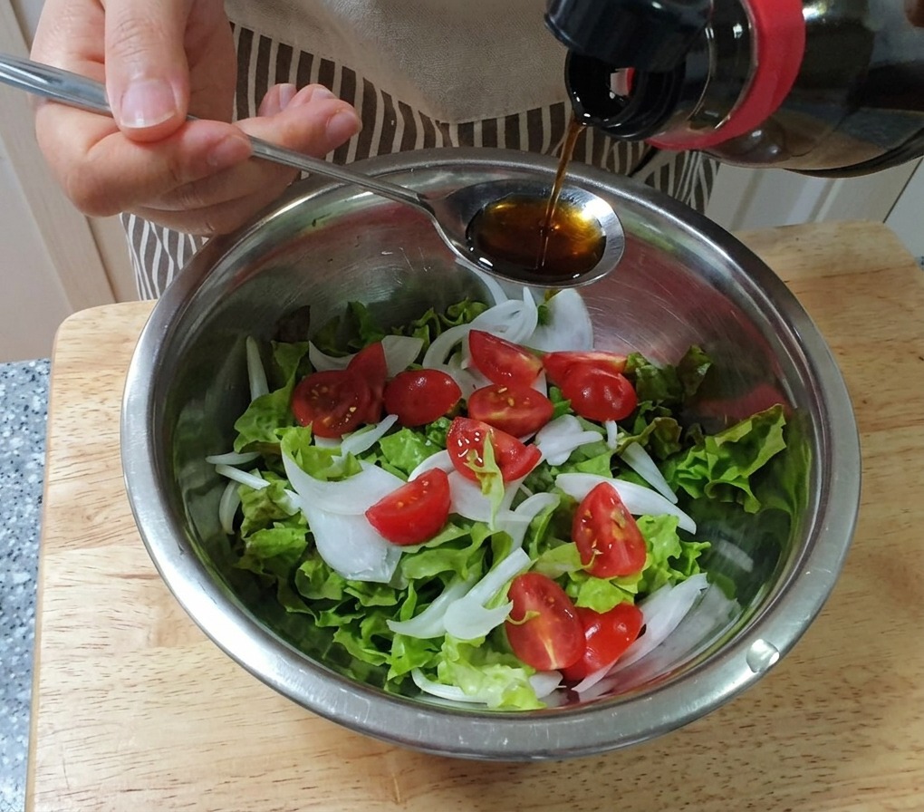 Dressing being poured from a spoon over a bowl of lettuce, onions, and cherry tomatoes.