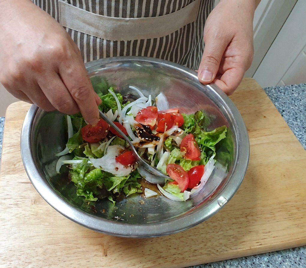 A hand mixing a fresh vegetable salad with a spoon in a large stainless steel bowl.