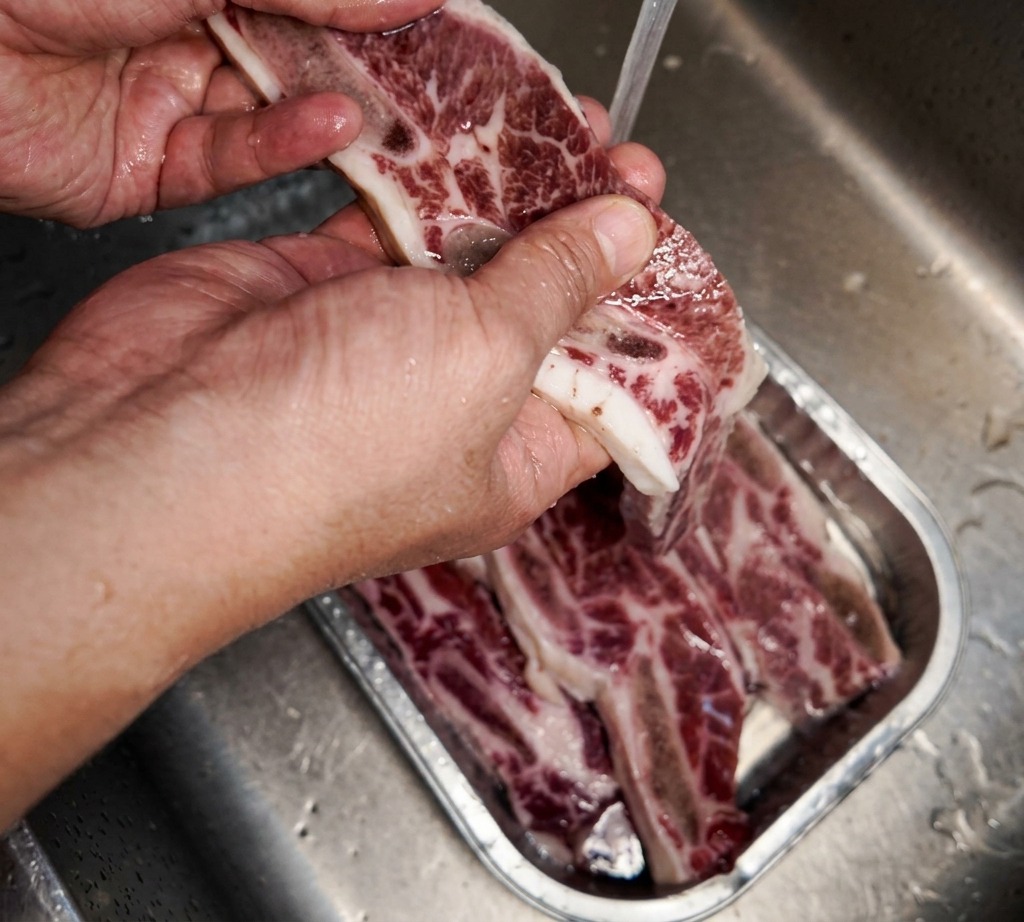 A hand holding a piece of marbled beef short rib under a stream of cold tap water over a stainless steel sink.