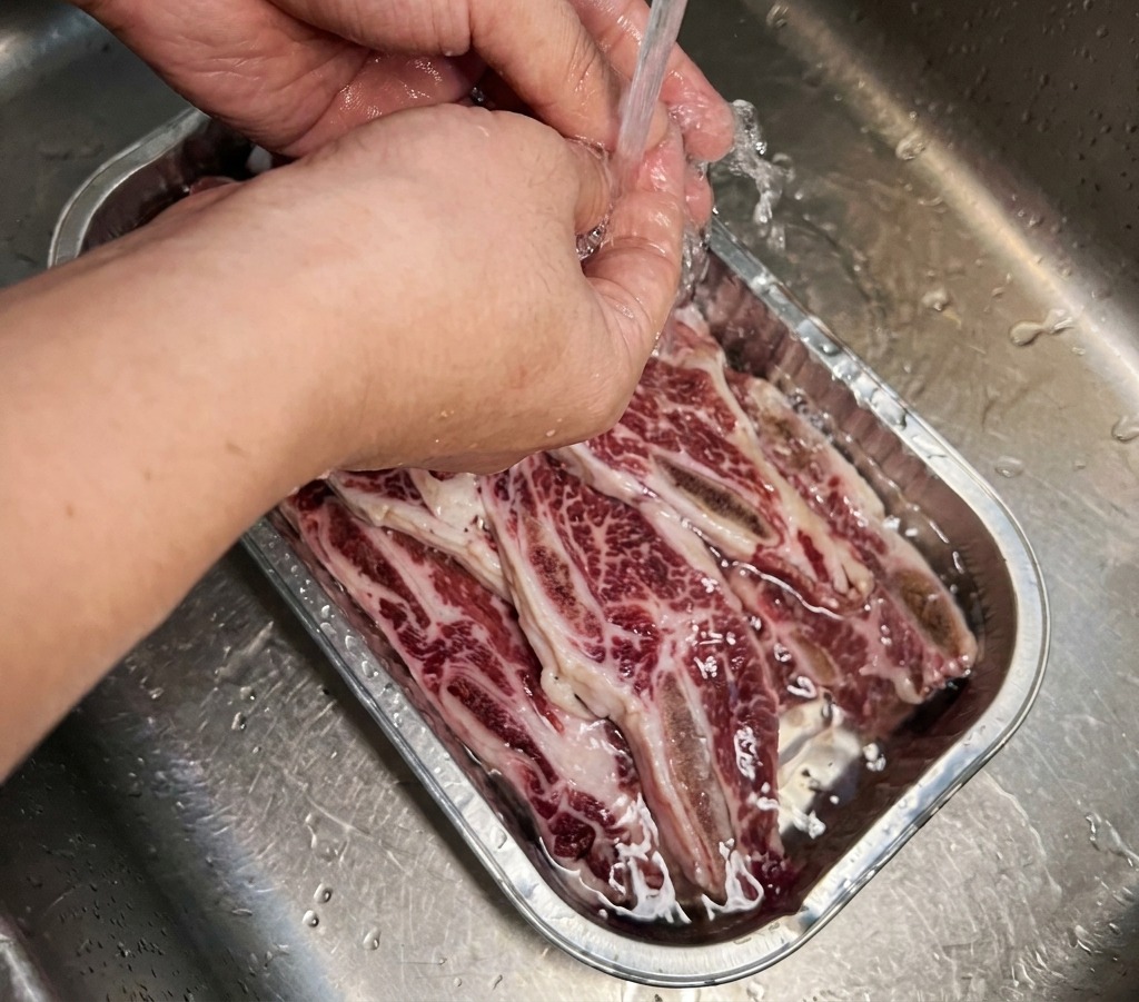 Beef short ribs submerged in a metal tray being filled with clear cold water from a faucet.