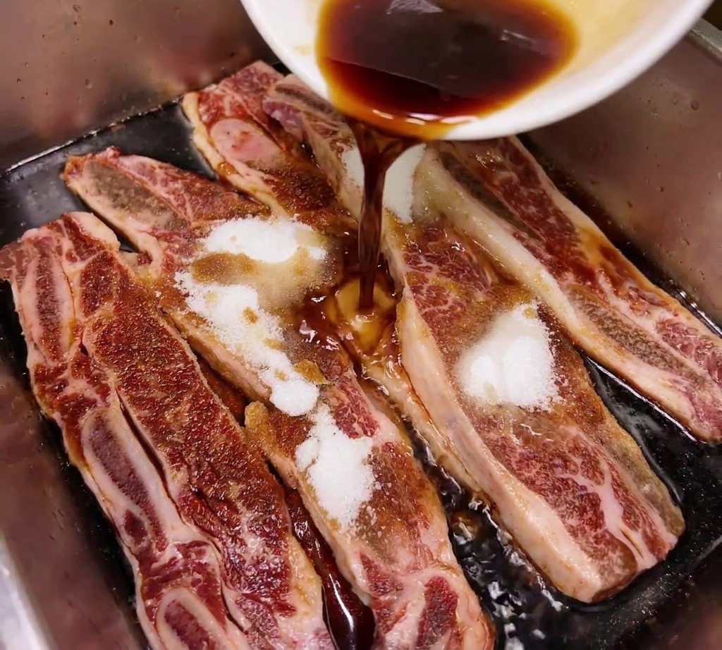Dark soy sauce or mirin being poured from a white bowl onto raw beef short ribs that have been sprinkled with white sugar in a metal tray.