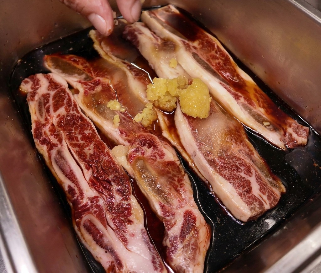 A hand placing a small mound of freshly minced ginger onto marinating beef short ribs in a stainless steel tray.