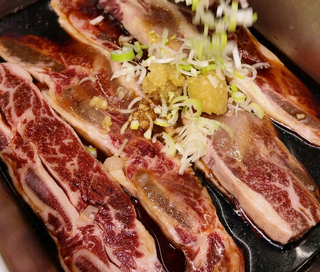 Chopped green onions being sprinkled by hand over a tray of raw beef short ribs already seasoned with garlic and ginger.