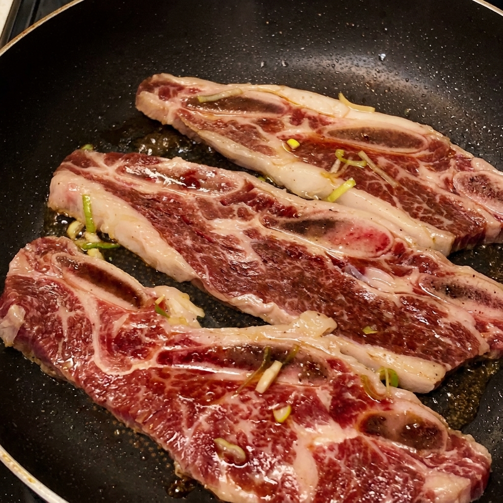 Three beef short ribs sizzling in a pan with the dark marinade bubbling and reducing into a thick, glossy glaze.
