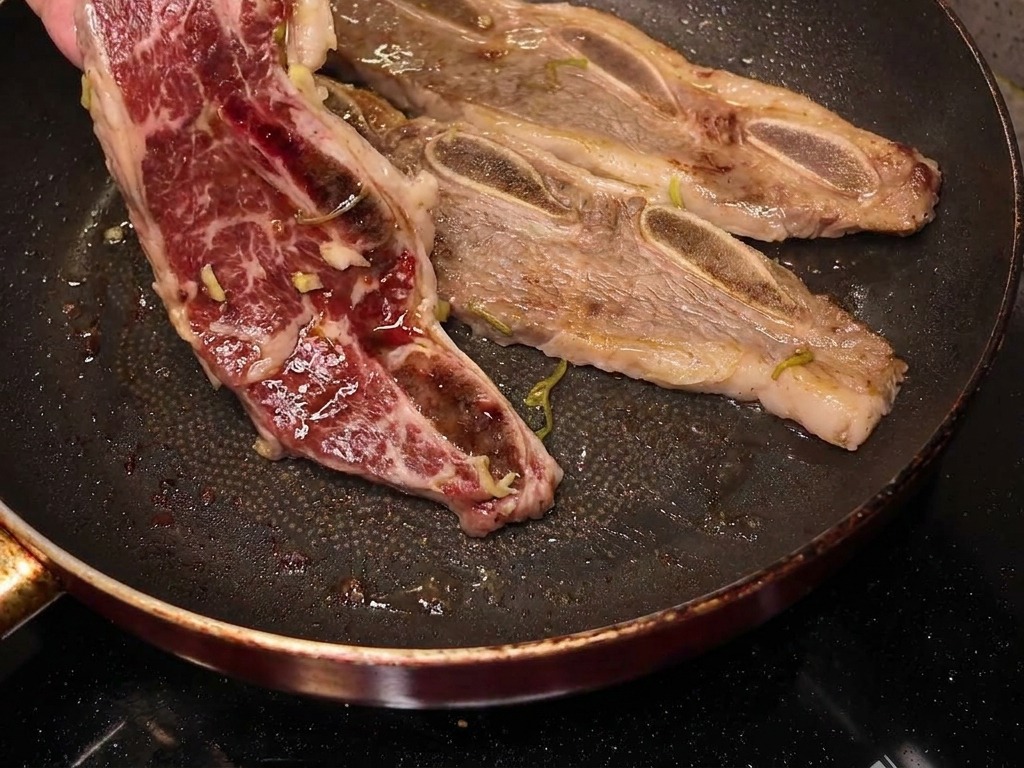 A hand flipping a partially cooked beef short rib in a frying pan to reveal a caramelized and browned surface.