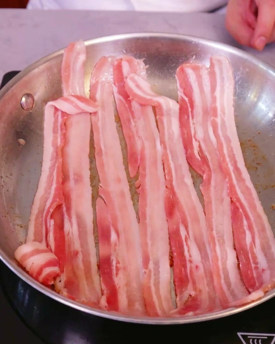 Slices of fresh, raw pork belly arranged neatly in a large stainless steel skillet on an induction stovetop.