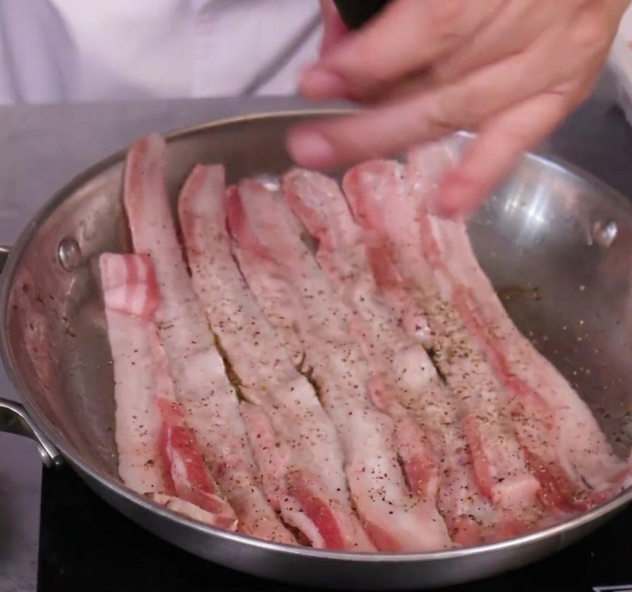 Slices of raw pork belly being seasoned with a manual black pepper grinder in a stainless steel skillet.