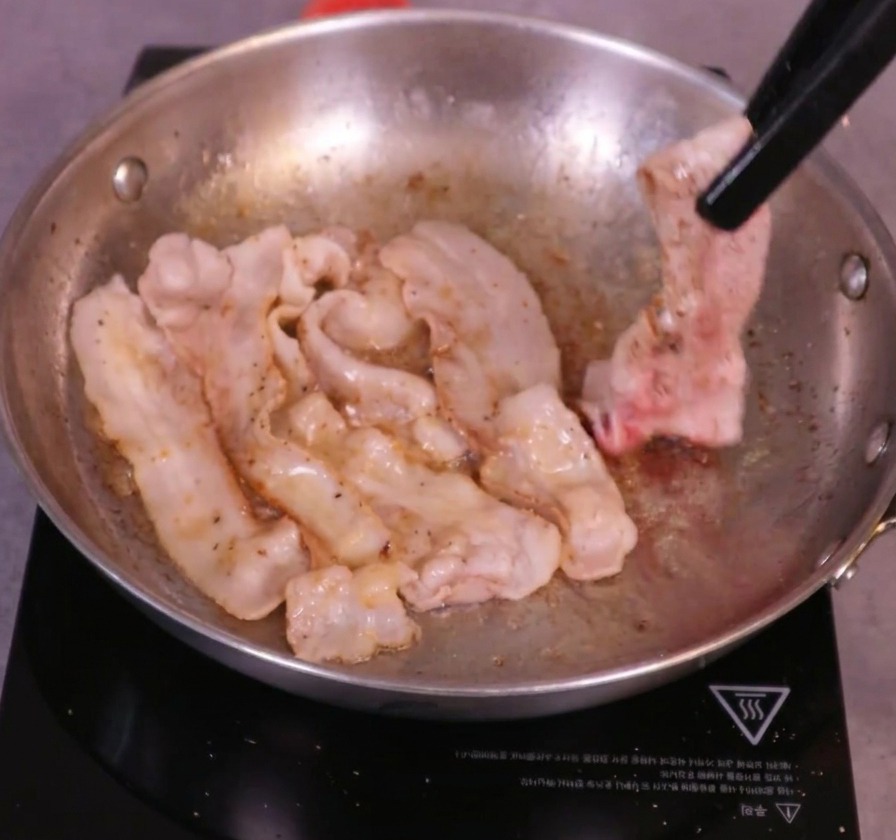 Golden-brown pork belly slices being turned over in a sizzling pan using black kitchen tongs.