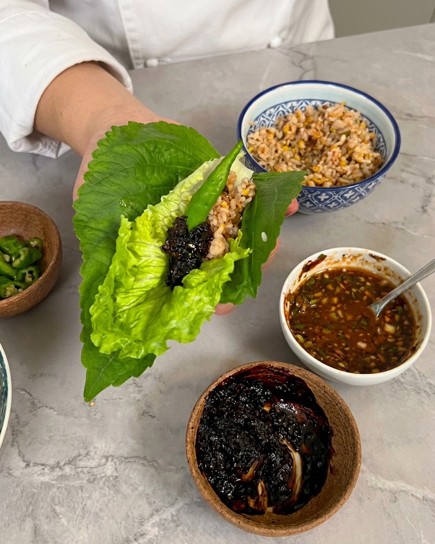 A hand holding a lettuce and perilla leaf wrap topped with a spoonful of dark barbecue sauce and a green chili pepper.