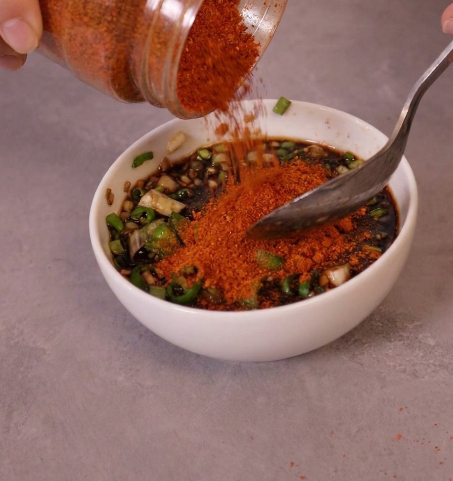 Bright red fine chili powder being poured from a jar into a bowl of prepared sauce base and green chilies.