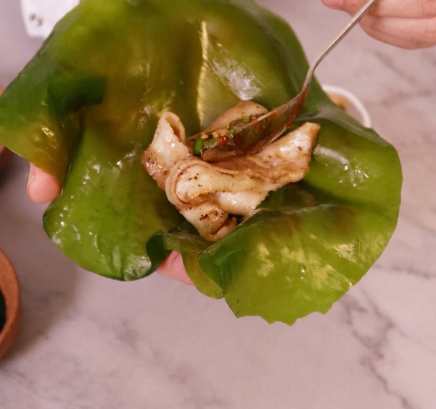 A hand holding a large, translucent green kelp leaf with a piece of cooked pork belly placed in the middle.