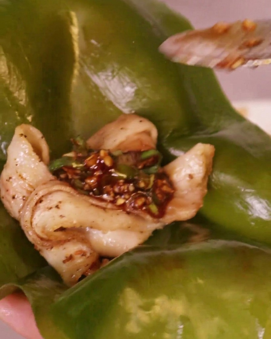 Spicy red chili sauce being dolloped onto a piece of grilled pork belly resting on a green kelp leaf.