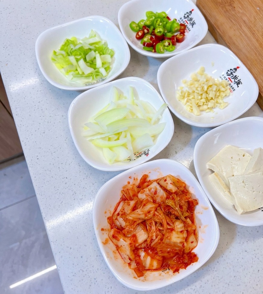 A collection of small white bowls containing chopped scallions, green and red peppers, minced garlic, sliced onions, tofu, and kimchi.