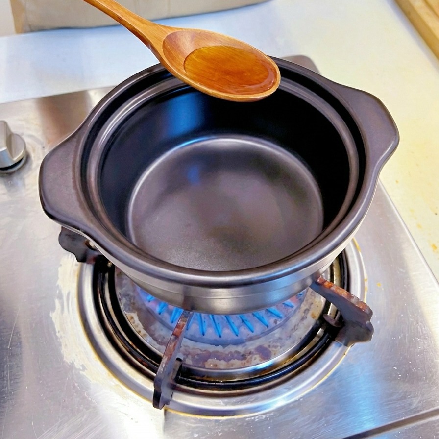 A wooden spoon pouring golden sesame oil into a black stone cooking pot on a gas stove.