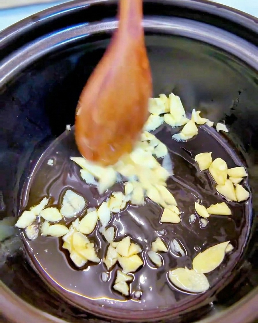 Minced garlic being stirred in sesame oil at the bottom of a black pot using a wooden spoon.