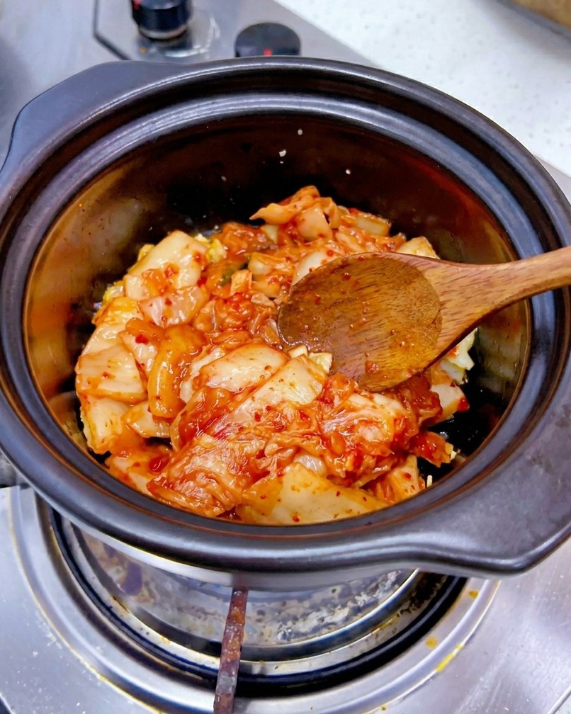 A portion of chopped red cabbage kimchi being added to a pot with sautéed garlic.