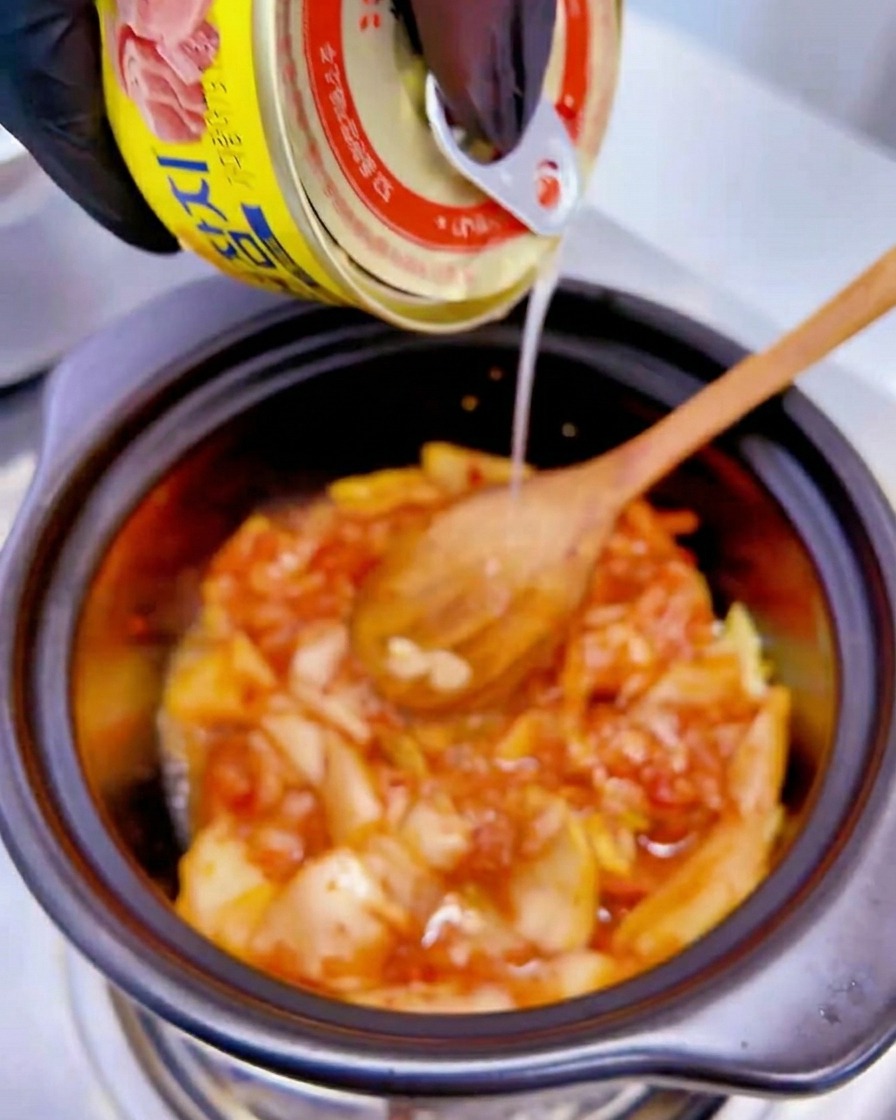 Liquid from a yellow can of tuna being poured over the kimchi mixture in the pot.