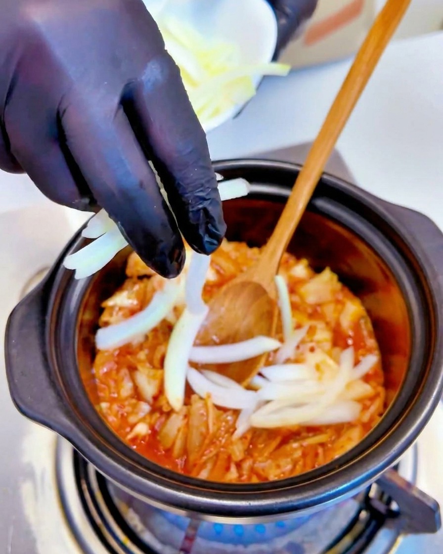 Sliced white onions being added to the simmering kimchi mixture in a pot by a hand wearing a black glove.