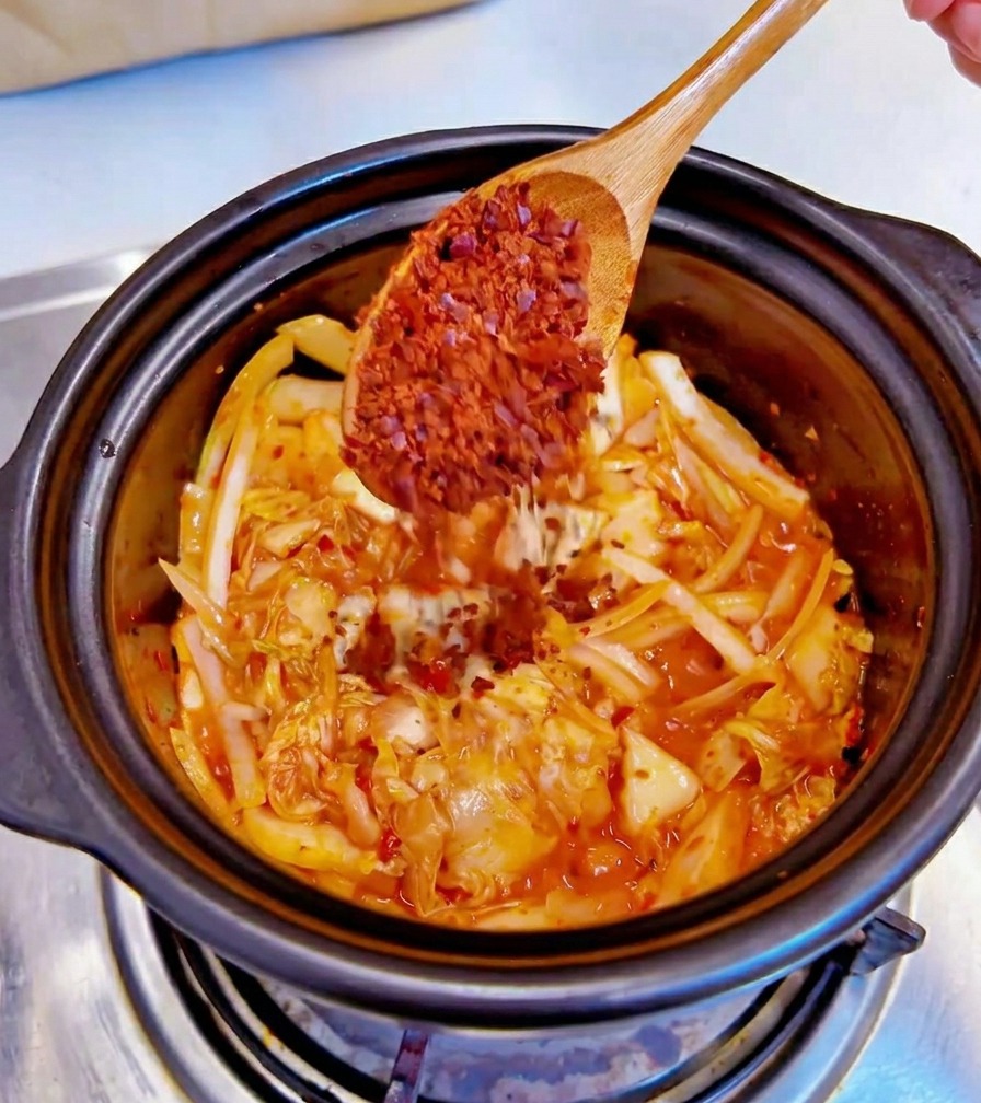 A wooden spoon adding coarse red chili flakes into a black pot containing sautéed kimchi and sliced onions.