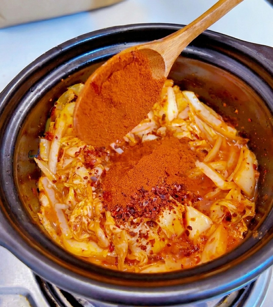 Fine red chili powder being added from a wooden spoon onto the seasoned kimchi and onion mixture.