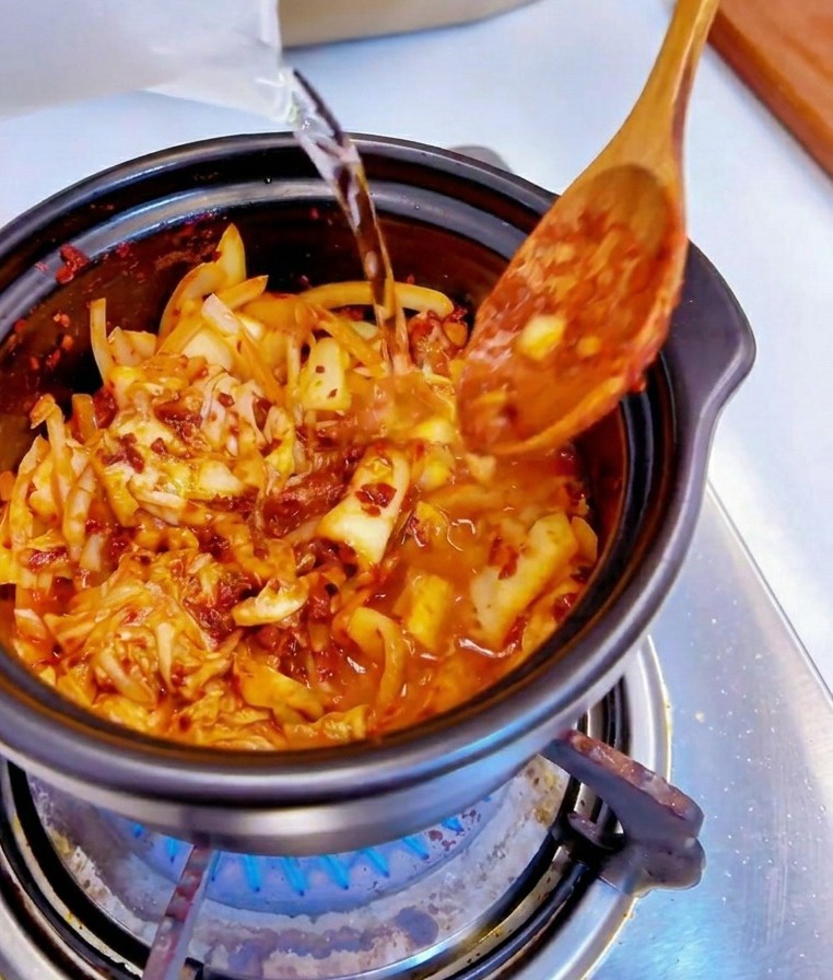Water being poured from a clear plastic measuring cup into a black pot filled with spicy kimchi and onions.
