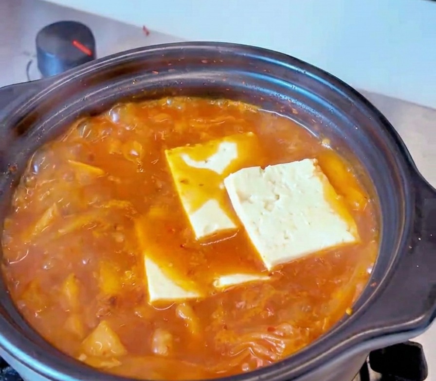 Rectangular slices of white tofu resting in a bubbling red kimchi stew broth.