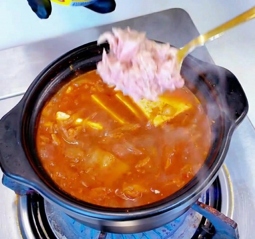 Chunks of light pink canned tuna being spooned into the middle of a simmering red stew.