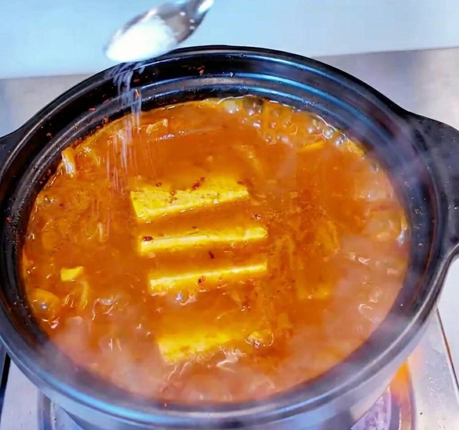 A small metal spoon sprinkling white salt over the surface of a bubbling tuna kimchi stew.