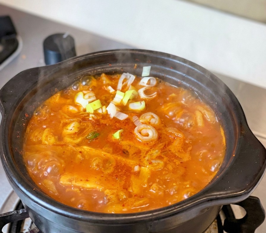 A bubbling red Tuna Kimchi Stew in a traditional black clay pot on a stove, topped with fresh green scallions and pepper slices.