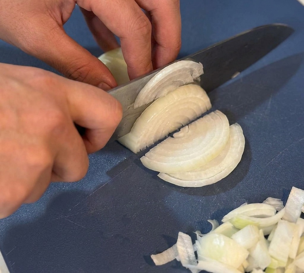 Hands using a chef knife to thinly slice a white onion on a blue cutting board.