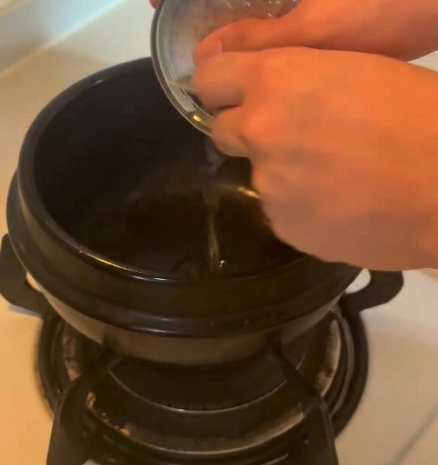 Savory oil being poured from a metallic tuna can into a dark ceramic pot on a stovetop.