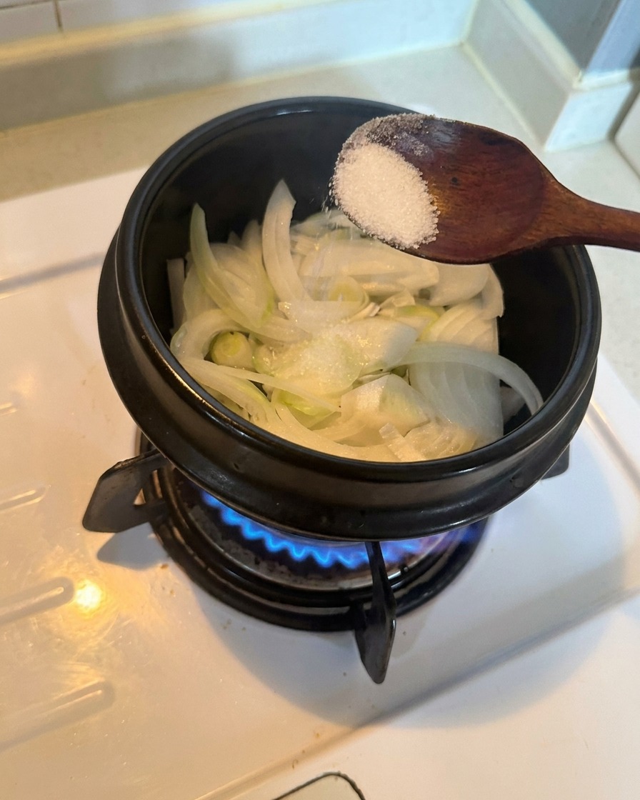 Sliced white onions in a pot being seasoned with a spoonful of white sugar over a lit gas burner.