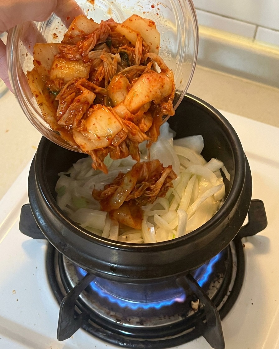 Adding a bowl of vibrant red chopped kimchi into a pot containing sautéed onions and scallions.