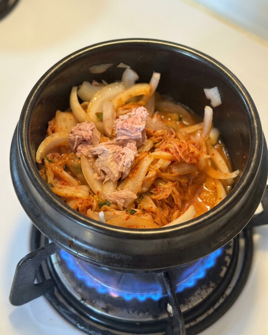 Chunks of canned tuna sitting on top of a bed of sautéed kimchi and onions in a black pot.