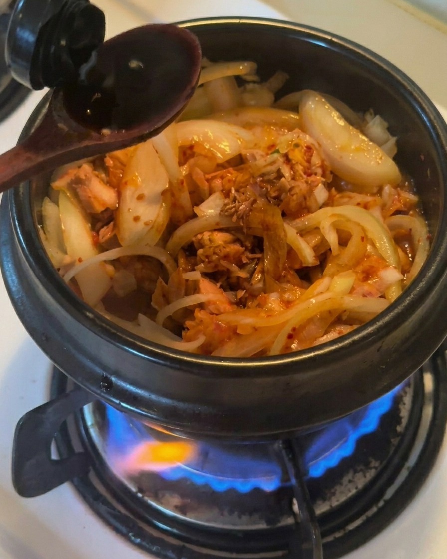 A wooden spoon pouring dark soy sauce into a black earthenware pot containing stir-fried onions, tuna, and kimchi.