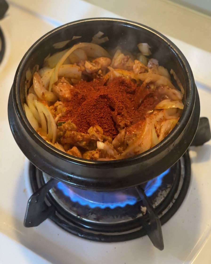 A large mound of bright red gochugaru sitting on top of sautéed kimchi and onions in a black stone pot over a gas flame.