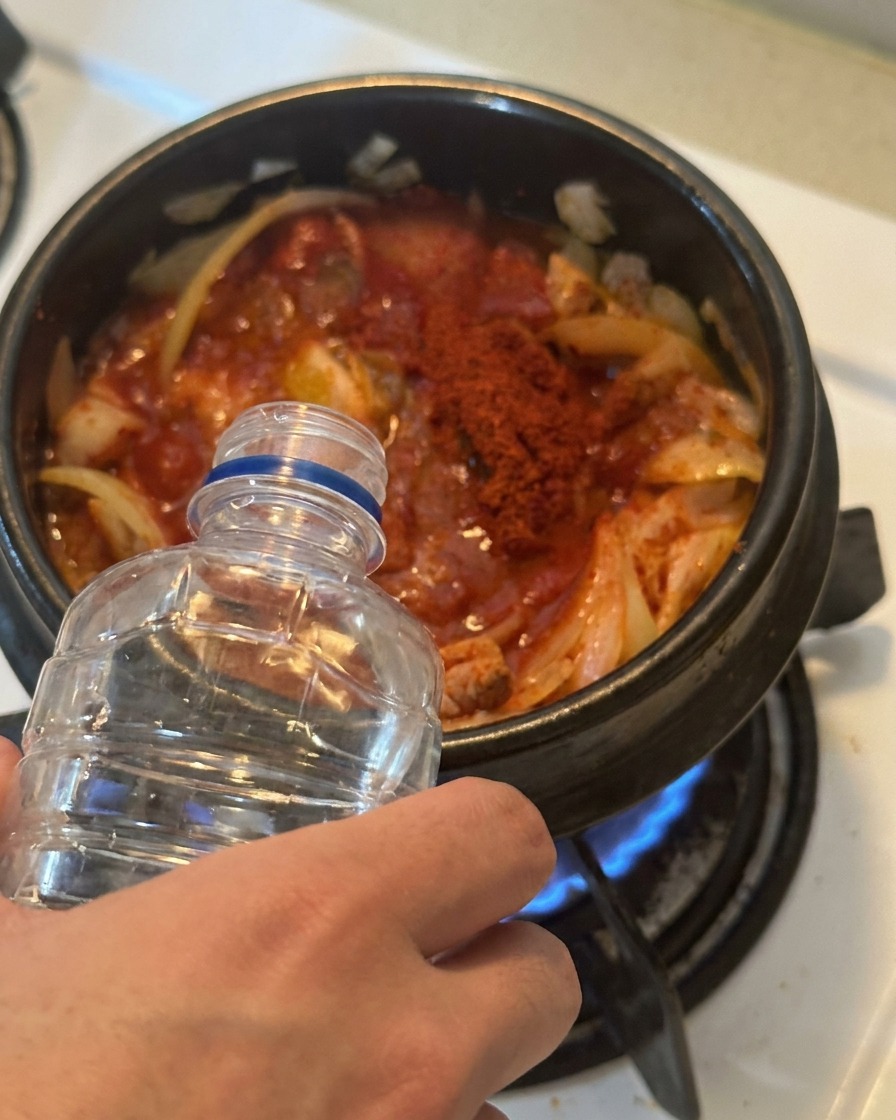 Water being poured from a clear plastic bottle into a pot filled with red-seasoned kimchi and tuna.