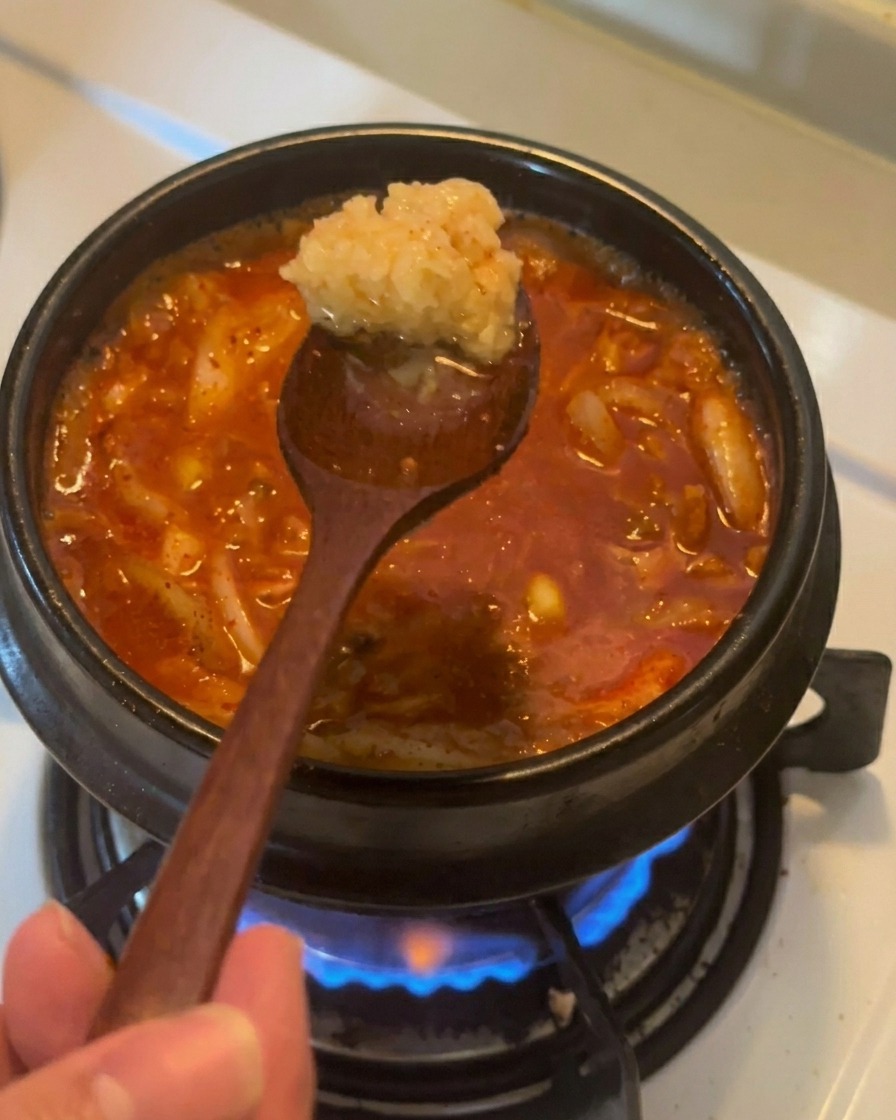 A wooden spoon holding a scoop of minced garlic over a bubbling red tuna kimchi stew.